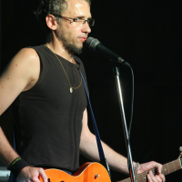 A man with curly hair, glasses, and a beard stands on stage, speaking into a microphone. He is wearing a sleeveless black shirt and a necklace. Holding an orange hollow-body electric guitar with a blue strap, he exudes Laba daba vibes. The dark background emphasizes the stage lighting on the performer.