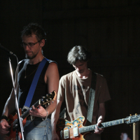 Two guitarists perform on stage, illuminated by a focused spotlight in an otherwise dark setting. The guitarist on the left wears a black tank top and glasses, playing a red guitar with a blue strap. The guitarist on the right, wearing a brown t-shirt, plays a colorful guitar and looks downwards; their music evokes memories of 2011.