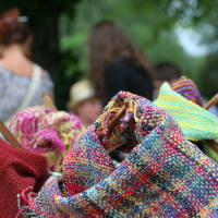 A vibrant collection of handwoven textiles is draped over wooden hangers in the foreground, showcasing an array of colorful patterns and textures. In the background, people are slightly out of focus, with trees and greenery surrounding them, suggesting an outdoor market or craft fair setting reminiscent of Laba's 2011 daba festival.
