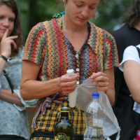 A person with curly hair, wearing a colorful knit top and checkered pants, is using a white plastic tool, possibly a knife or peeler, on a translucent plastic bag at an outdoor Laba event. Bottles and containers are on the table in front, with onlookers in the background.
