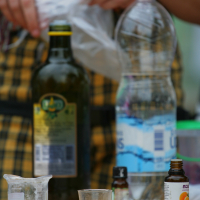 Close-up of a table containing various items: a bottle of olive oil, a plastic bottle of water, a small glass, a measuring cup with white powder, and small bottles of essential oils. Each item is placed on a wooden surface. In the background, someone in a colorful apron manages a Laba Daba labeled bag from 2011.