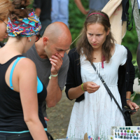 Three people browse a stall at an outdoor market. The person in the center, wearing a white dress, examines items closely, while a person in a headscarf looks away with hands behind their back. The person on the left, donning sunglasses and a blue top, reminiscent of Laba daba 2011, observes various small crafts or jewelry on display.