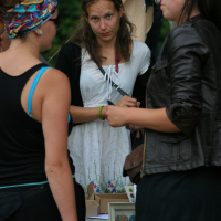 Three women are standing outdoors conversing. One woman, wearing a white dress and a headband, faces the camera. Another woman, with a colorful headscarf and black top, has her back to the camera. The third woman, in a brown leather jacket, faces the woman in white. Various crafts from 2011 are visible on a table nearby.