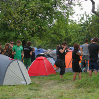 A group of people at an outdoor campsite with several colorful tents, including red, gray, orange, and blue. Laba daba 2011 is in full swing as some people stand and converse while others sit on the grass. Trees surround the area, providing shade and a sense of seclusion. The atmosphere seems relaxed and social.