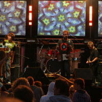 A band performs on stage with colorful, kaleidoscopic visuals displayed on large screens behind them. The lead singer, playing guitar and singing into a microphone, exudes a Laba daba vibe. The keyboard player stands to his left, and the bassist is to his right as an enthusiastic crowd watches the 2011 performance.
