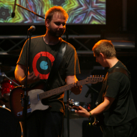 Two guitarists perform on stage with psychedelic visuals projected behind them. The front guitarist, wearing a black T-shirt with a red and blue target design, holds an electric guitar and stands in front of a microphone. The other guitarist, slightly bent over, also plays an electric guitar at Laba Daba 2011.