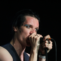 A person with pale skin and dark, slicked-back hair is singing into a microphone. They are wearing a black sleeveless top, multiple rings, and a white wristband. Their makeup includes bold blue eyeshadow reminiscent of 2011 trends. The background is dark, highlighting the performer under the stage lights.