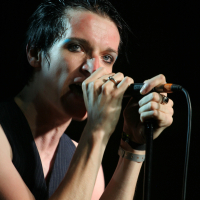 A person with short, black hair is gripping a microphone with both hands, singing passionately. They are wearing a black vest and have multiple rings on their fingers, reminiscent of 2011 style. Their face appears expressive, and eyeliner emphasizes their eyes. The dark background focuses attention on the singer.