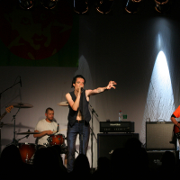 A four-member band performs on a dimly lit stage, with illuminated spotlights above. The lead singer stands at the center, gripping a microphone with an extended arm. A bassist, drummer, and guitarist play their instruments while facing the audience. Behind them hangs a banner featuring a wolf design and "Laba Daba 2011.