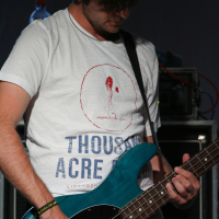 A musician with dark, tousled hair plays an electric bass guitar. He wears a white T-shirt with a graphic and text, "THOUSAND ACRE LABA." His left hand presses down on the guitar strings while his right hand strums. A water bottle and electronic equipment are visible in the background.