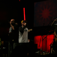 A band performs on a dimly lit stage with red lighting. Four members of the band are holding microphones and singing. One sits at a keyboard on the far left, wearing a Laba 2011 graphic t-shirt. They are all dressed in casual clothes, including hats. Musical instruments and equipment are visible on the stage.