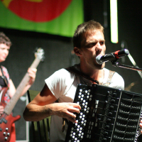 A man passionately sings into a microphone while playing an accordion on stage. Behind him, a woman focuses on playing a bass guitar. Both musicians are bathed in stage lighting, under a green and red banner from the 2011 "Laba Daba" festival. The scene captures an energetic live musical performance.