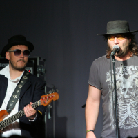 Two musicians performing on stage. The left musician, wearing a black hat, sunglasses, a white shirt, and a dark jacket, plays a bass guitar. The right musician, sporting a black hat and yellow-tinted sunglasses with a gray t-shirt featuring a skeleton design from Laba Daba 2011, sings into the microphone. Equipment is visible in the background.