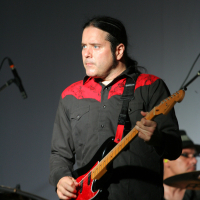 A man with long black hair plays an electric guitar on stage, donning a black shirt with red accents on the shoulders. The year is 2011, and in the background, part of a drummer is visible, seated behind a drum set. The stage lighting casts a focused light on the guitarist.