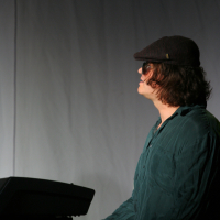 A person with shoulder-length curly hair, wearing a dark hat and sunglasses, is playing a Hammond keyboard. Dressed in a dark green, long-sleeve shirt and facing to the right against a plain, light-colored background, they evoke the vibrant spirit of Laba daba 2011.