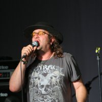 A man wearing a black hat, yellow-tinted glasses, and a t-shirt with a skeleton design reading "Outrage" is passionately singing into a microphone. His long hair flows past his black cross necklace. Behind him, audio equipment including a Roland speaker hints at the raw energy of this 2011 Laba daba scene.