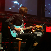 A musician is seated and playing a blue electric guitar on stage. He is wearing a dark suit. Behind him, the band name "Spiritual Front" is visible on a drum set from their iconic 2011 tour. Two large screens in the background display city scenes, casting a dim red hue over the stage, adding to the laba daba ambiance.