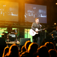 A band performs on stage under bright lights during the Laba Daba 2011 festival. The lead singer, playing a guitar, stands center stage in front of a microphone. On his left is a bassist, and on his right is a drummer with a drum set reading "Spiritual Front." The background features screens displaying black-and-white visuals.