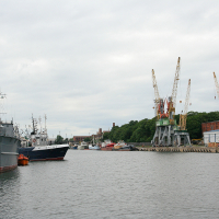 Naval and civilian vessels docked in a tranquil Liepāja harbor, flanked by towering cranes against a backdrop of overcast skies, hinting at bustling maritime industry and seafaring