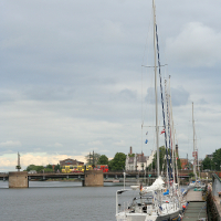 A row of sailboats moored at a dock in Liepāja on a cloudy day, with calm water reflecting a bridge and buildings in the distance.
