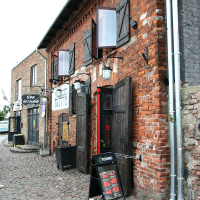 A rustic red brick building with wooden doors, hosting a bar and restaurant in Liepāja, on a cobblestone street; a blackboard menu and empty crates sit out front under clear skies