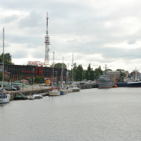 Cloudy skies over a bustling harbor in Liepāja, with an array of boats and ships moored along the quay; waterfront buildings and a towering communication mast stand in the background, suggesting