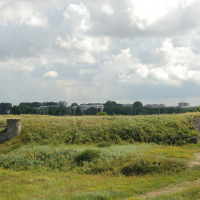 Verdant fields under a partly cloudy sky with remnants of old military bunkers near Liepāja, foregrounding a modern city skyline, illustrating a contrast between history, nature, and urban development