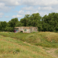 A weathered, graffiti-covered concrete building sits abandoned amidst verdant, grassy mounds on a sunny day with scattered clouds. This secluded structure in Liepāja, Latvia is surrounded by tall, lush green trees, and a narrow dirt path winds through the grass towards it, enhancing the scene's secluded feel.