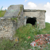 An abandoned concrete bunker in Liepāja, partially covered with graffiti, sits in a field with overgrown vegetation and a pile of rubble in the foreground under a partly cloudy sky.