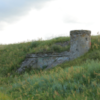 An old concrete structure in Liepāja, possibly a bunker, emerges from a grass-covered hillside, bearing signs of graffiti, under a cloudy sky, evoking a sense of forgotten history amidst