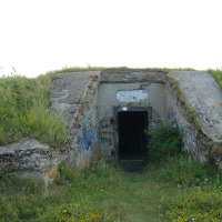 An abandoned concrete bunker with graffiti-covered walls emerges from a grass-covered mound in Liepāja, its dark entryway offering a silent testament to its mysterious past.