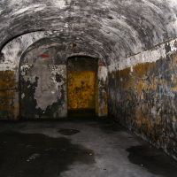A dimly lit, abandoned curved tunnel in Liepāja with peeling, graffiti-marked walls leads to a weathered yellow door, creating an atmosphere of neglect and mystery.
