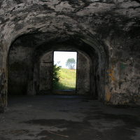 An old, arched stone tunnel with chipped paint and visible wear in Liepāja leads towards an outdoor exit, revealing a glimpse of greenery and a clear sky.