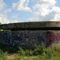 A weathered World War II concrete bunker covered in colorful graffiti, sitting under a cloudy sky in a grassy field near Liepāja, symbolizing a contrast between historical conflict and modern expression.