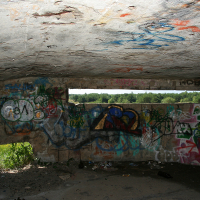 Graffiti-covered walls under a concrete structure in Liepāja, with colorful tags and drawings, offer a glimpse into local urban art and expression, contrasted by the bright, natural daylight peek