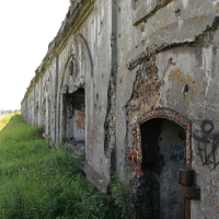 A long, weathered stone wall in Liepāja with large, arched openings in various stages of decay. The wall is covered in cracks and patches of missing plaster. Graffiti is visible on some sections. Overgrown grass and wild plants flank the base of the wall, leading to an open grassy area in the background.