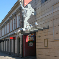 A dynamic metal sculpture of a guitarist extending from the facade of a building above a pedestrian street in Liepāja, showcasing a fusion of art and urban architecture under a clear blue sky.