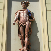 A bronze sculpture of a youthful, athletic male figure, assumedly an ancient hero or deity, is adorned with a draped cloth, set against a framed niche on a sunlit wall in Liepā
