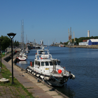 A sunny day at a calm port in Liepāja showing moored boats along a quay, with industrial buildings, cranes in the distance, and a clear blue sky overhead.