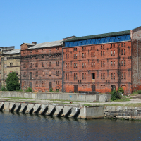 An imposing red brick industrial building, with signs of aging and disuse, stands by a calm waterway in Liepāja under a clear blue sky.