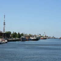 A clear day over the bustling port of Liepāja, with various ships docked along the quay and a blue sky reflecting in the calm water, while industrial cranes loom in the