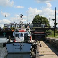A survey vessel named "aquasurveyor" is moored on a calm riverbank in Liepāja, beneath a blue sky dotted with clouds, with a bridge and greenery in the