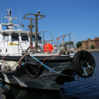 A tugboat with prominent fenders moored in a calm harbor in Liepāja, under clear blue skies, with urban buildings visible in the background.