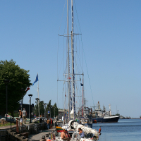 A sailboat with a red hull is docked at a waterfront promenade in Liepāja under clear blue skies, surrounded by calm waters and distant ships, inviting a day of maritime adventure.