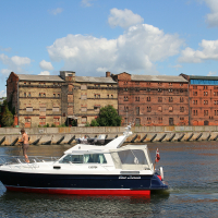 A luxury boat sails on a river in Liepāja, passing in front of historic brick warehouses under a blue sky dotted with clouds, illustrating a blend of recreational boating and industrial heritage.