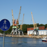 Sign indicating a pedestrian and bicycle lane in the foreground, with a waterfront and several cranes in the background. A small boat navigates the water near industrial buildings and trees under a clear blue sky, capturing a typical scene in Liepāja.