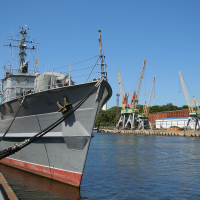 A military vessel moored at a calm dock in Liepāja, with towering cargo cranes in the background under a clear blue sky, signifying a blend of naval defense and commercial activity.