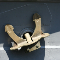 A shiny, gold-colored metal anchor is mounted on a dark grey surface with rivet details in Liepāja, suggesting a naval or maritime setting, with sunlight highlighting its contours.