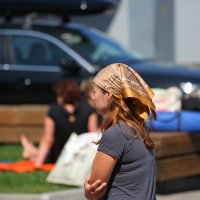 A person in a brown headscarf and grey top gestures with their hands while talking in Liepāja, with blurred figures sitting in the sunny background amidst parked cars.