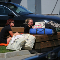 Two women sitting on the grass near a wooden ledge with several bags and a rolled-up sleeping bag in front of them. One woman with dark hair and sunglasses sits on an orange mat, while the other, with blonde hair, talks animatedly about their trip to Liepāja. A parked black car is visible in the background.